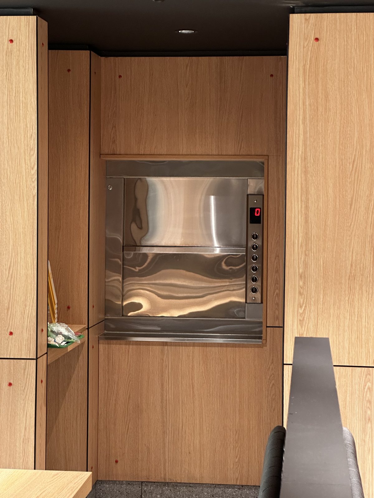 A small stainless steel dumbwaiter set into a wood-paneled wall, with buttons for floors 0 to 5